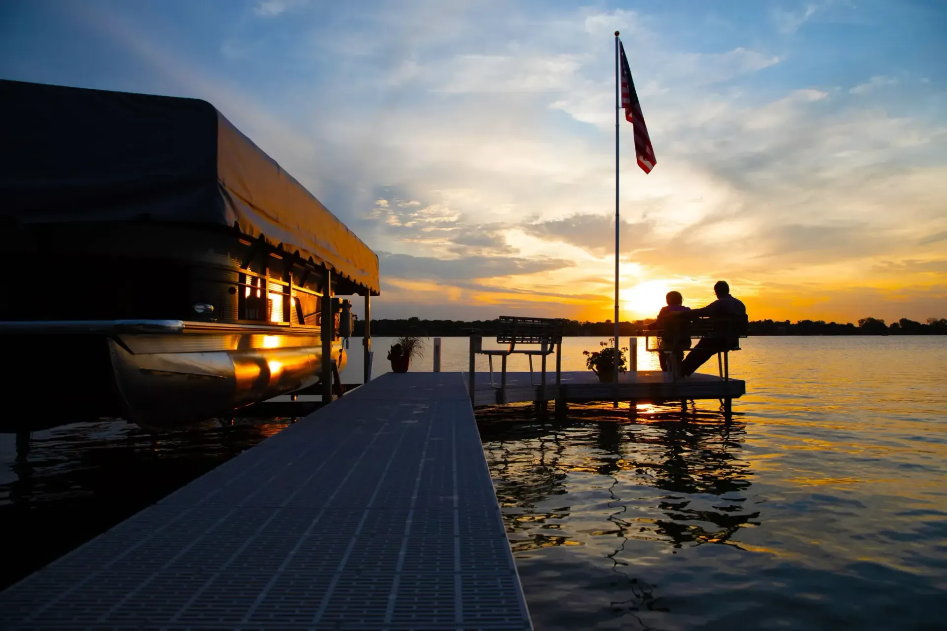 Dock with boat house and silhouetted figures watching sunset over water.