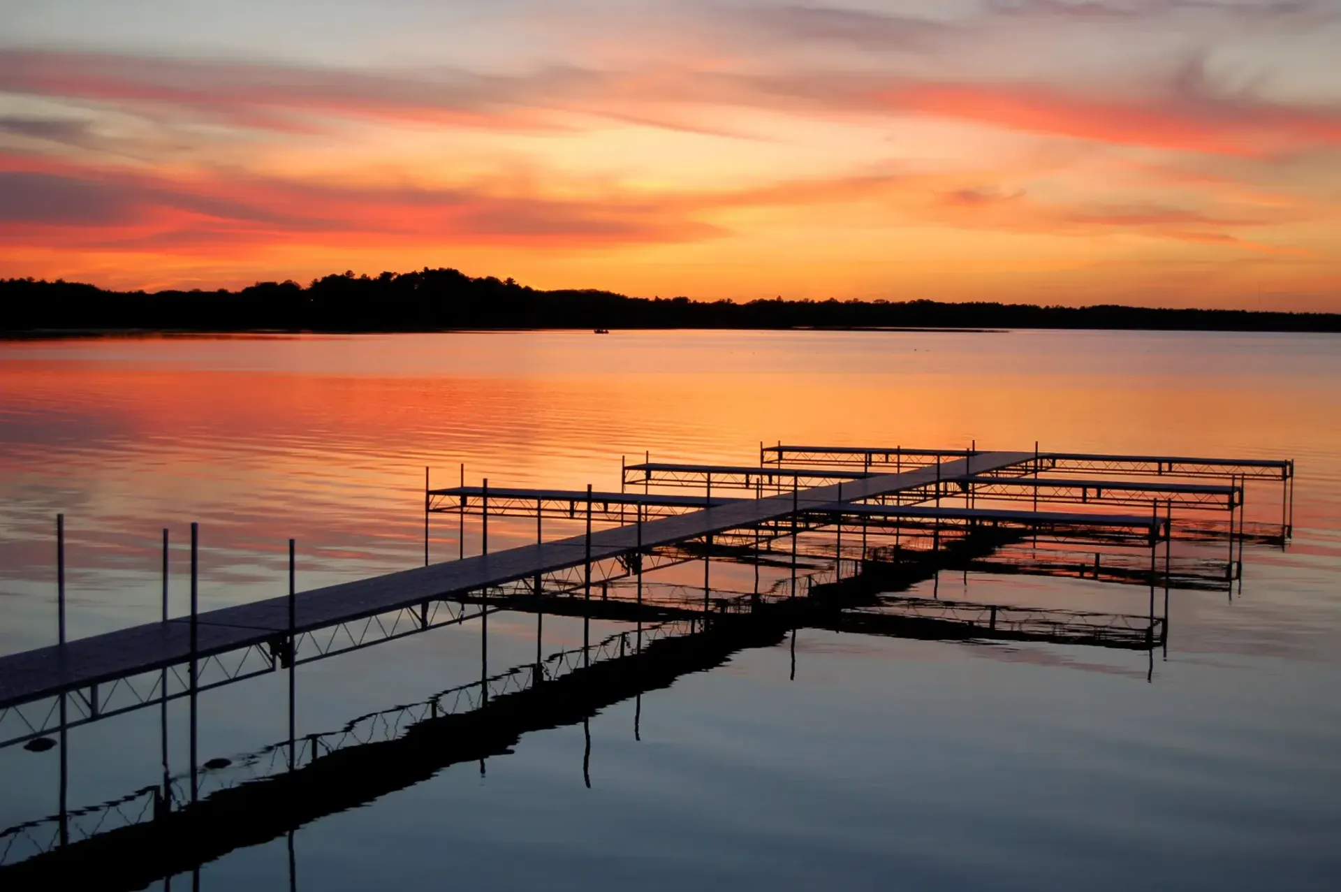 Sunset over calm lake, silhouetted dock extends into water reflecting colorful sky.