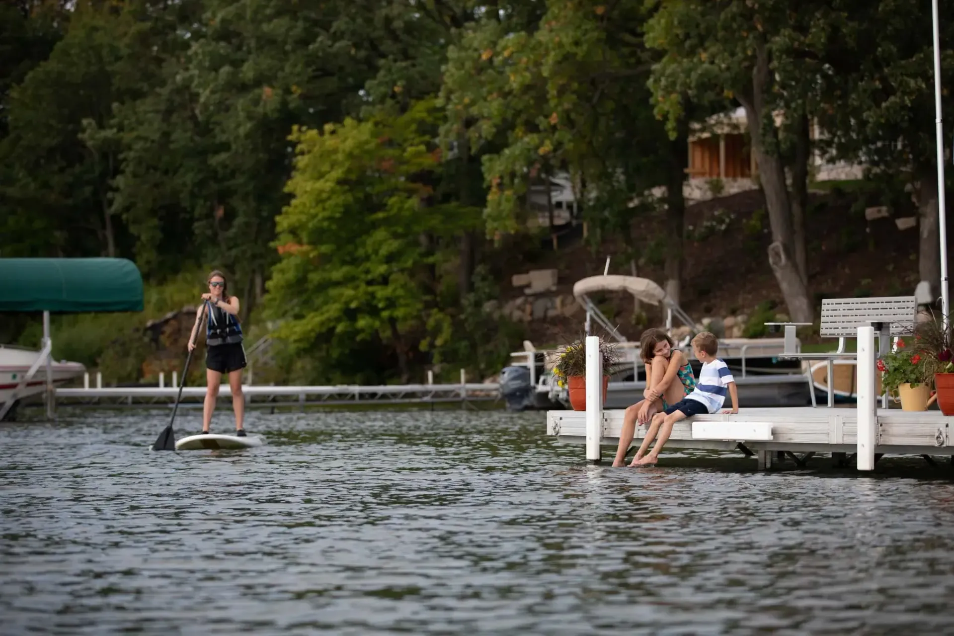 Woman paddleboards in a lake. Two people sit on a dock. Green trees and a house in the background.
