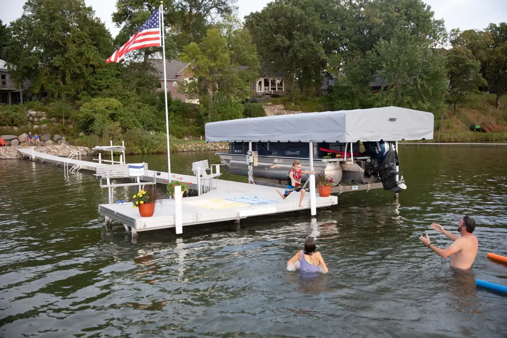 People swimming near a dock with a boat lift. American flag flies. Trees and houses in background.