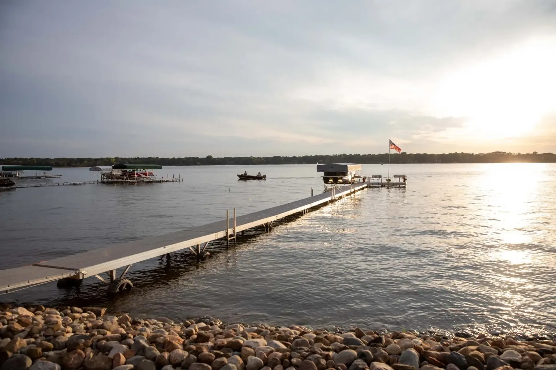 A wooden dock extends into a calm lake at sunset. Scattered boats and a flag in the distance.