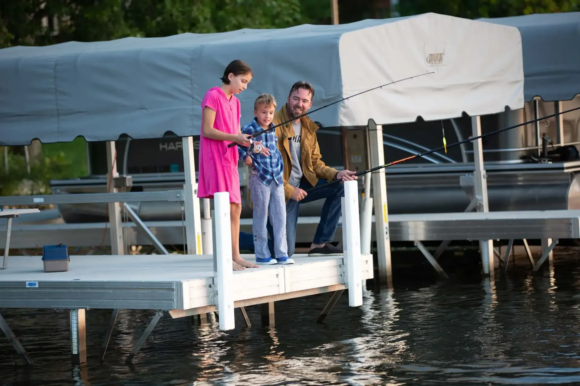 Family fishing from a dock: girl in pink, boy, and man with beard. Boats in background.