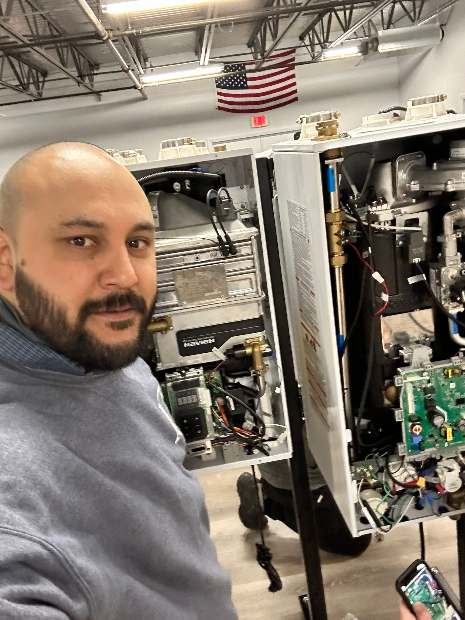 Man taking a selfie beside open electronic equipment in a workshop with an American flag overhead