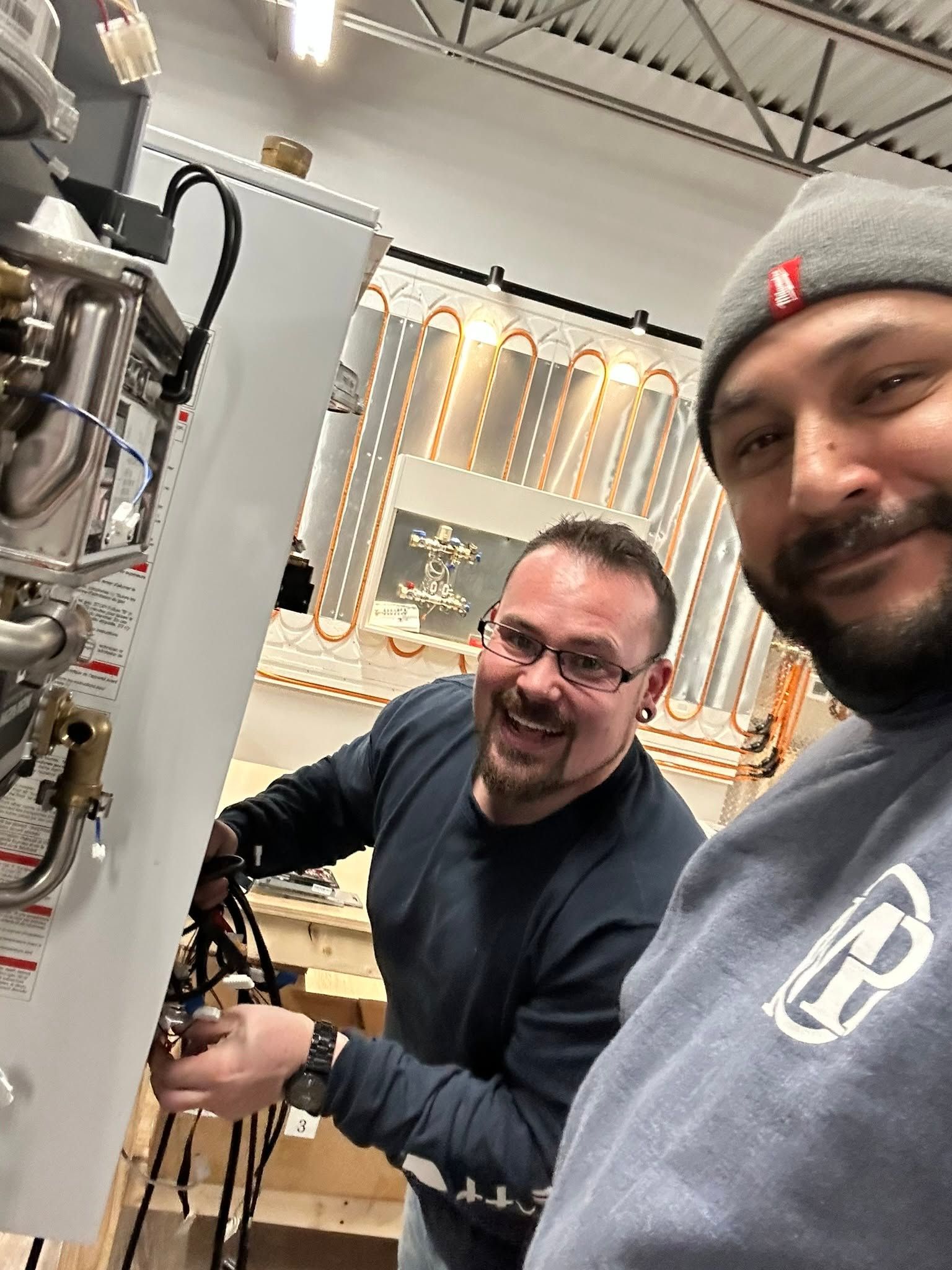 Two men smiling beside industrial machinery in a workshop, one wearing a gray beanie and blue shirt.
