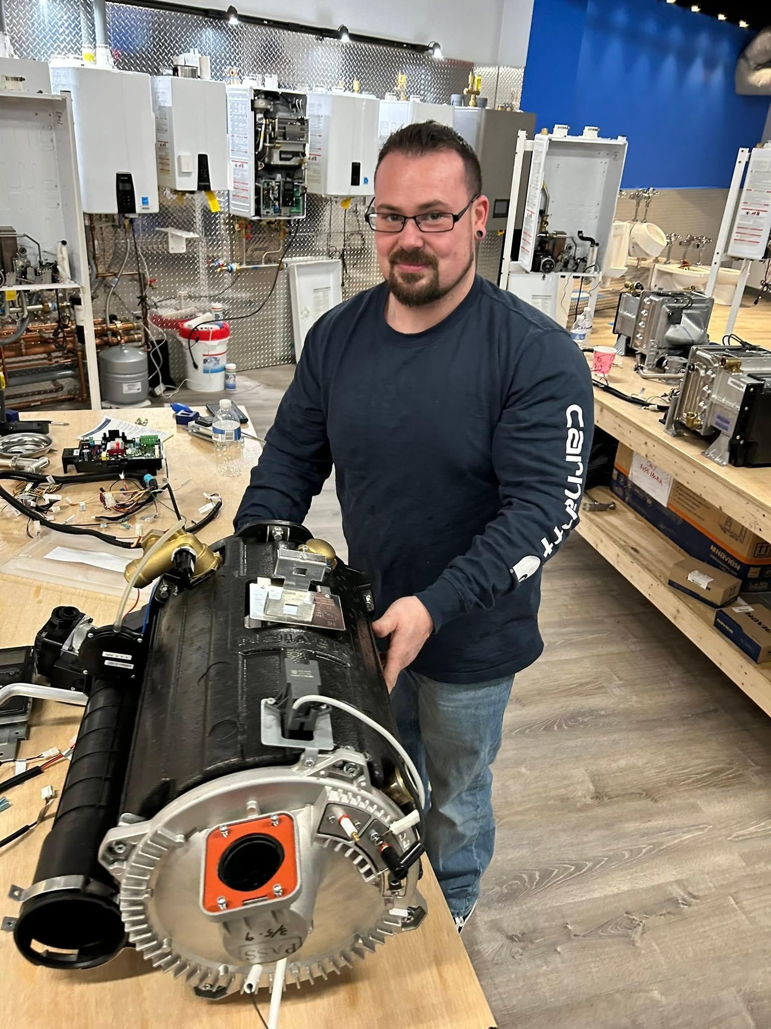 Man standing in a workshop holding a large engine component on a workbench.