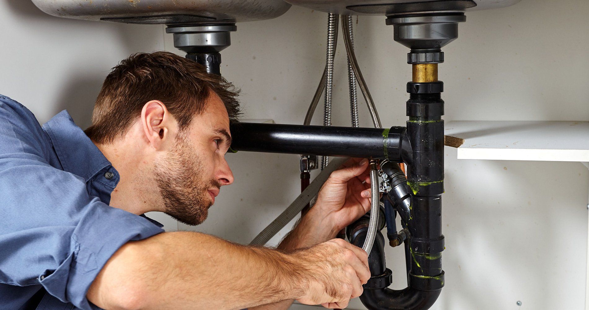 Plumber repairing a black under-sink pipe with a wrench in a kitchen cabinet