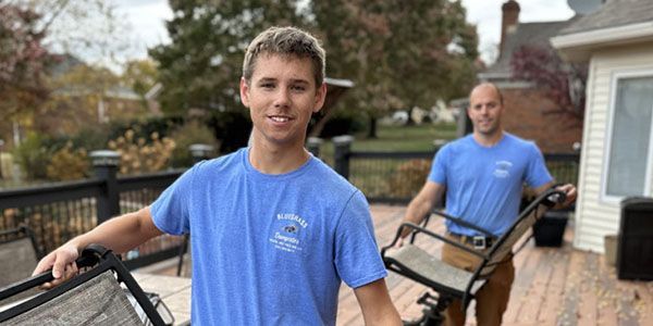 Two men in blue shirts are carrying chairs on a deck.