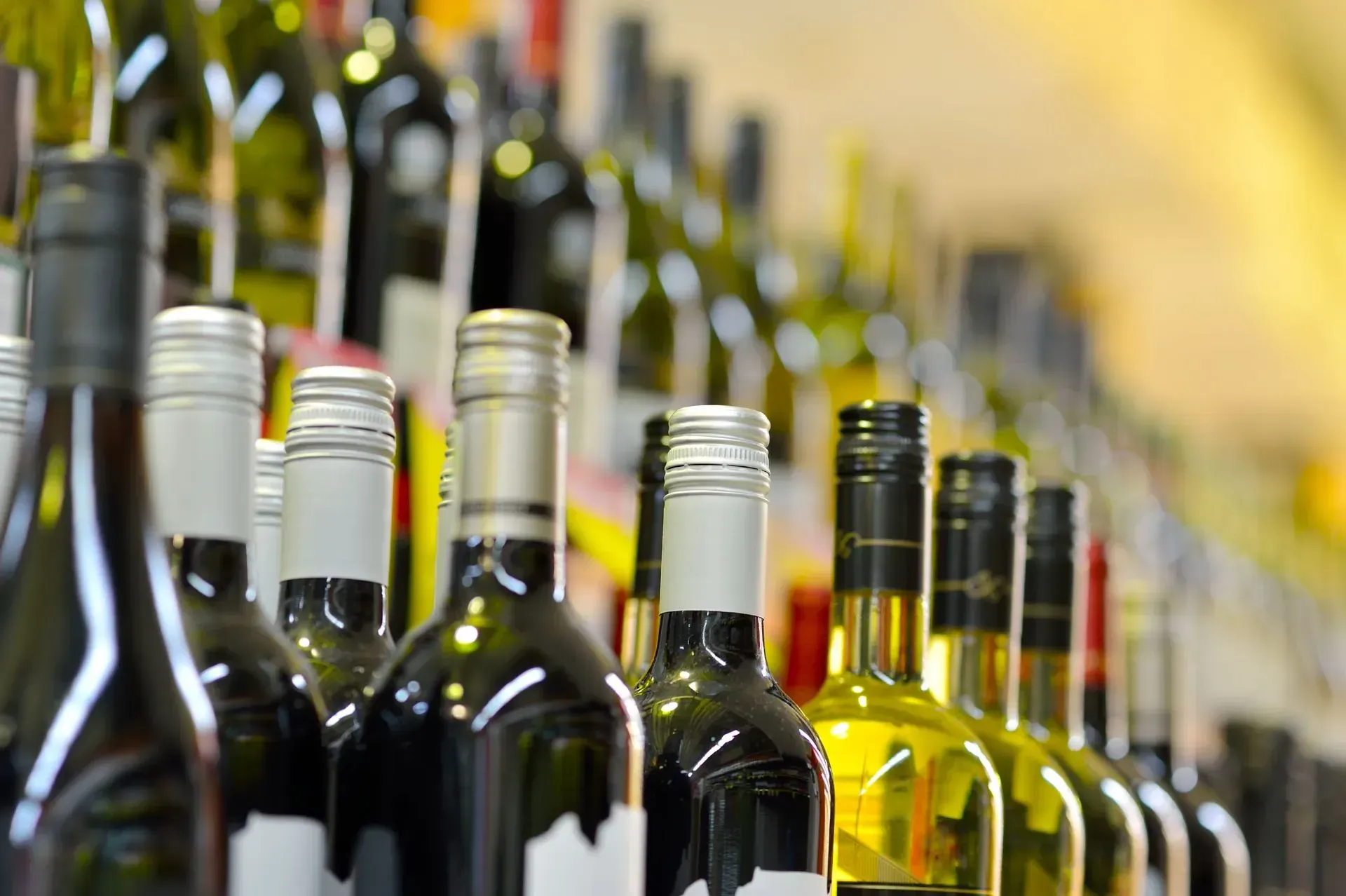 Rows of wine bottles with foil tops, some filled with red, some yellow-green, on a store shelf.
