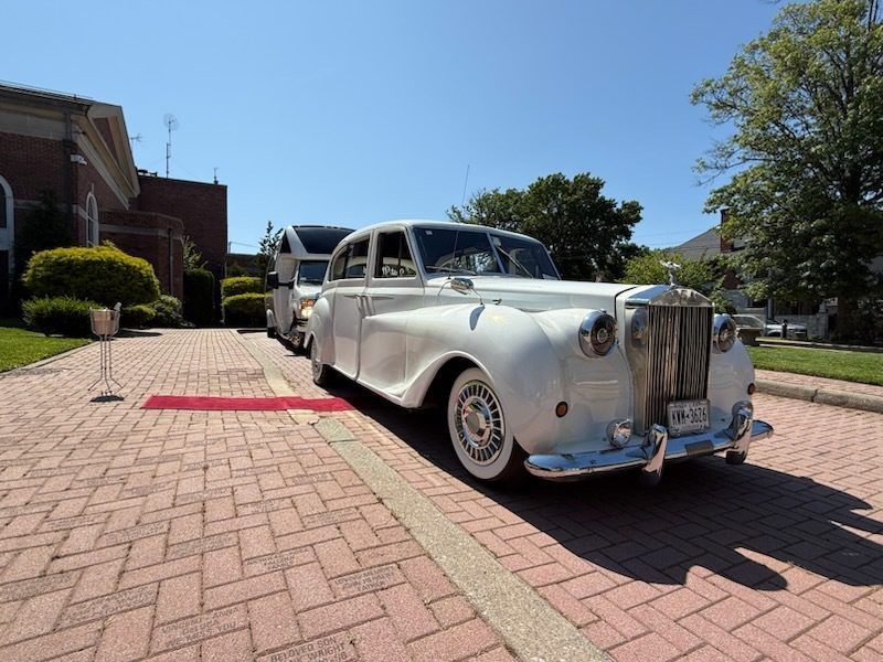 White vintage car parked on brick path with red carpet, near a building.