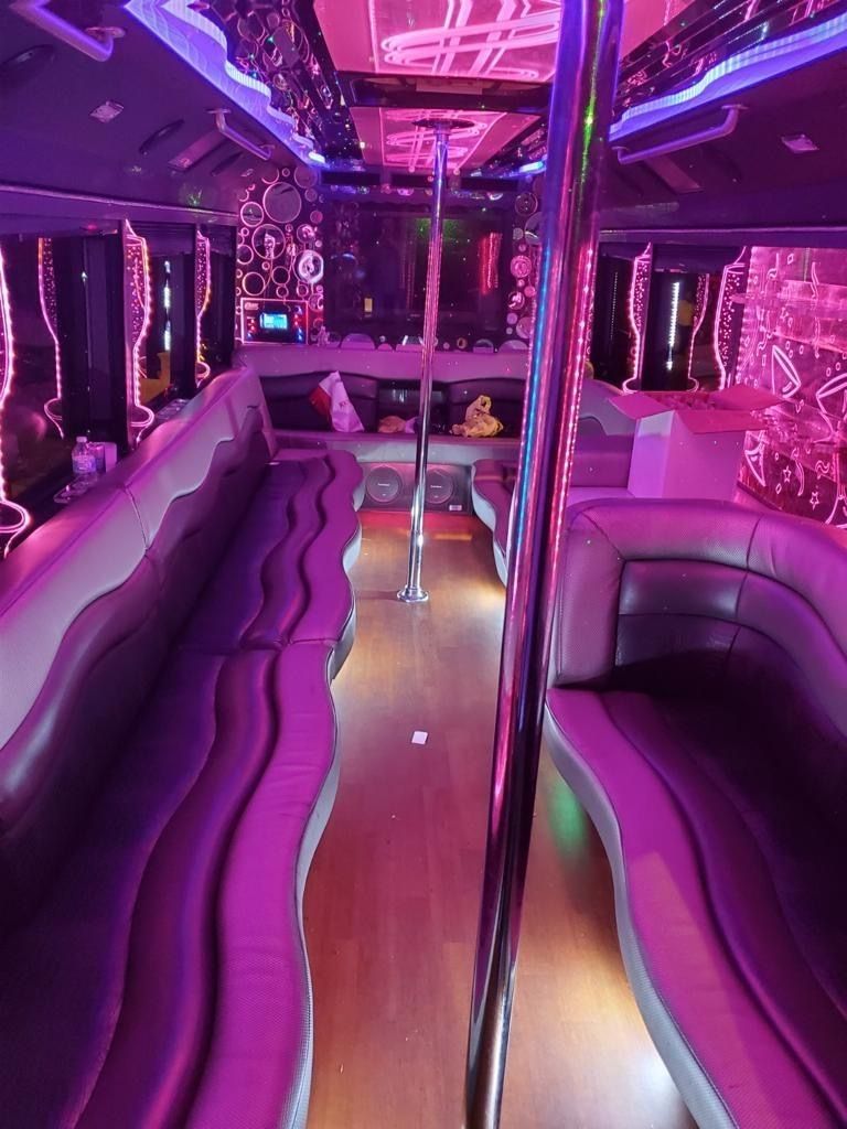 Interior of a party bus with purple lighting, two rows of seats, and a central pole.