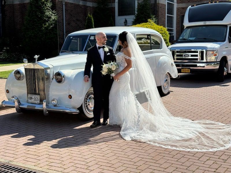 Newlyweds pose by a white vintage car. The bride wears a gown with a long veil, the groom, a tuxedo. Outside.