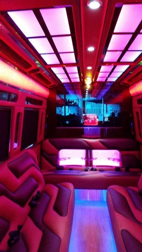 Interior of a party bus with pink, purple, and blue LED lights. Leather seating, a mirrored ceiling, and a wooden floor are visible.