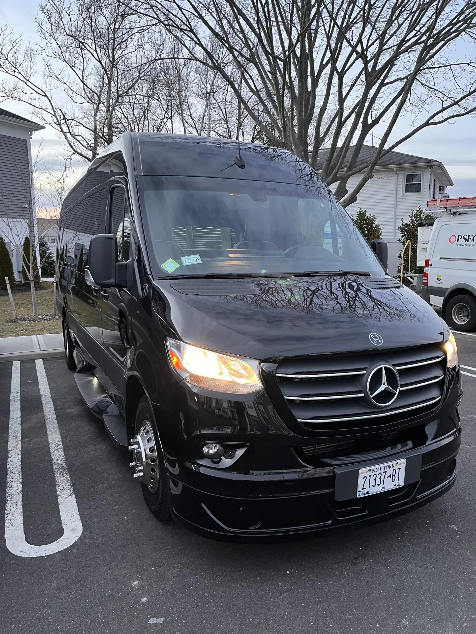 Black Mercedes-Benz Sprinter van parked in a lot, headlights on, near a building and trees.