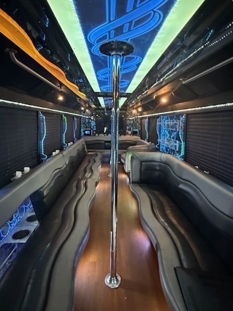 Interior of a party bus with black wraparound seating, a central pole, and colorful neon lights.