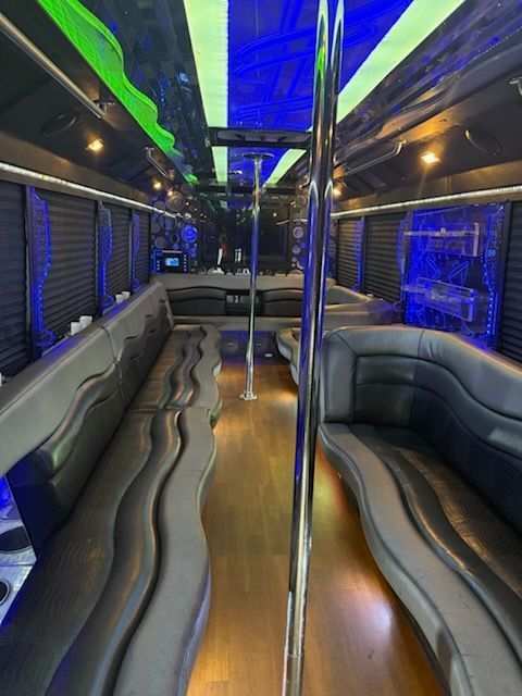 Interior view of a party bus with leather seating, a dance pole, and colorful overhead lighting.