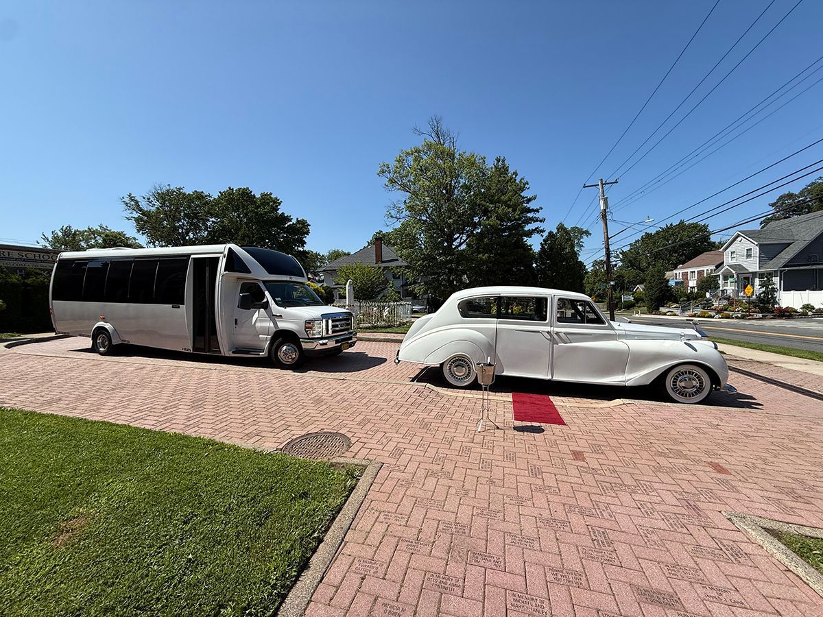 White bus and vintage car parked on brick driveway. Red carpet leading to the car.