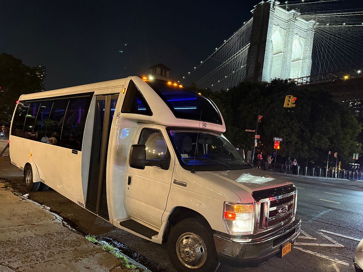 White passenger van parked near the Brooklyn Bridge at night, lights on.