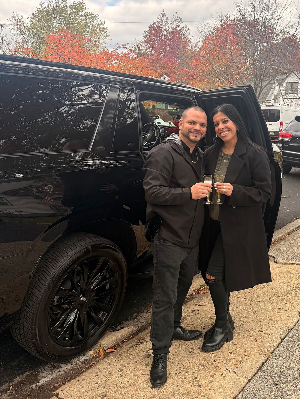 Couple toasting, smiling next to a black SUV with tinted windows. Fall foliage in the background.