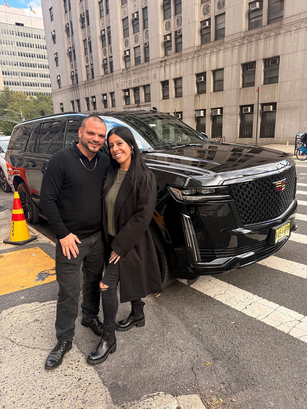 Couple standing next to a black Cadillac SUV on a city street.