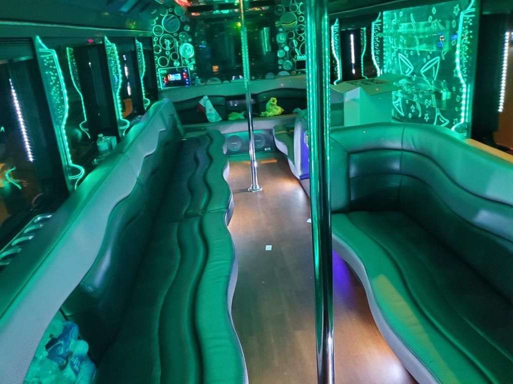Inside a party bus with green neon lights, seating, and a central pole.