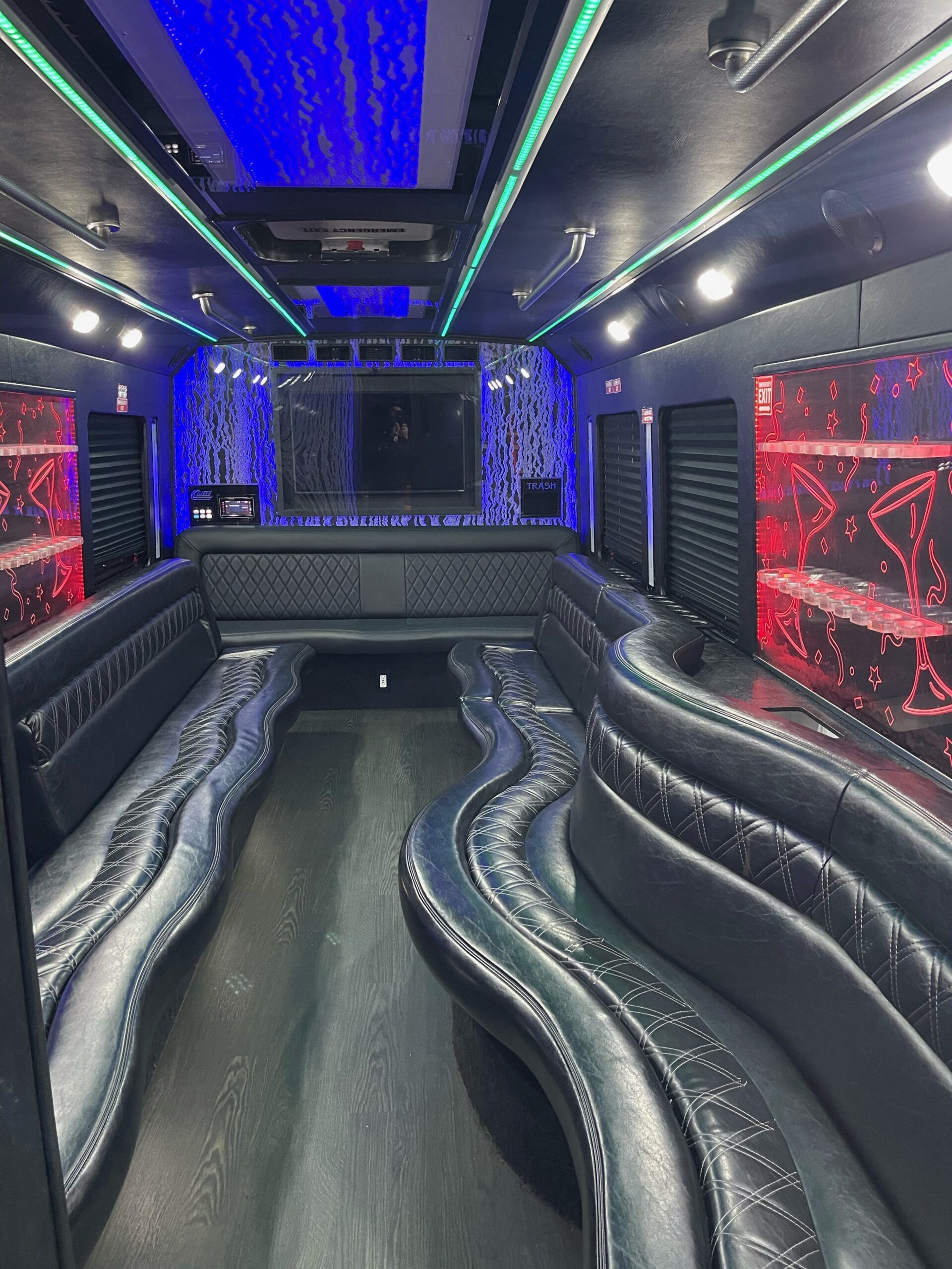 Interior of a party bus with black leather seating, colorful LED lights on the ceiling, and a bar area.