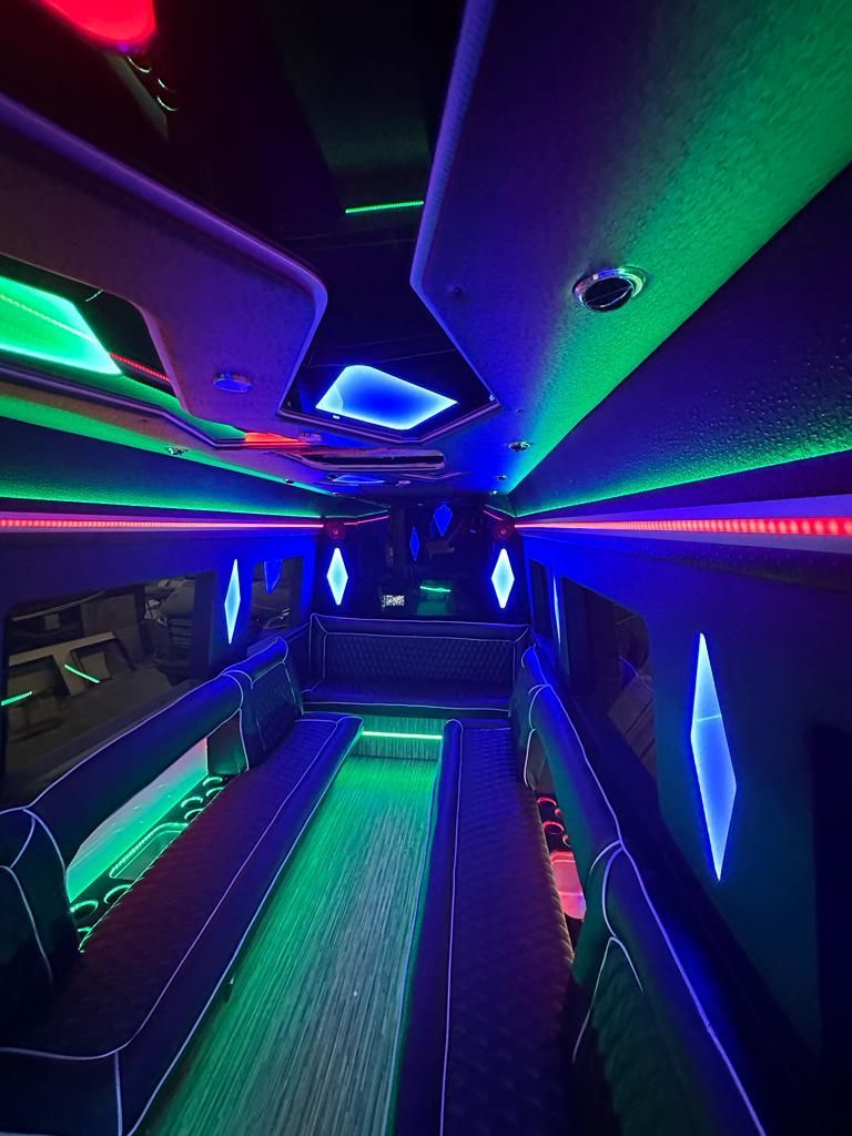 Interior of a party bus lit with green, blue, and red neon lights. Black seating lines the sides.
