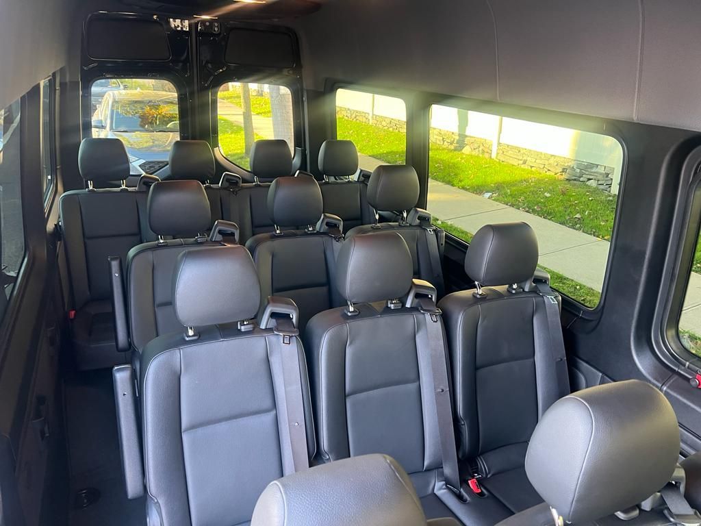 Interior of a black passenger van, featuring rows of black seats, windows, and bright outdoor light.