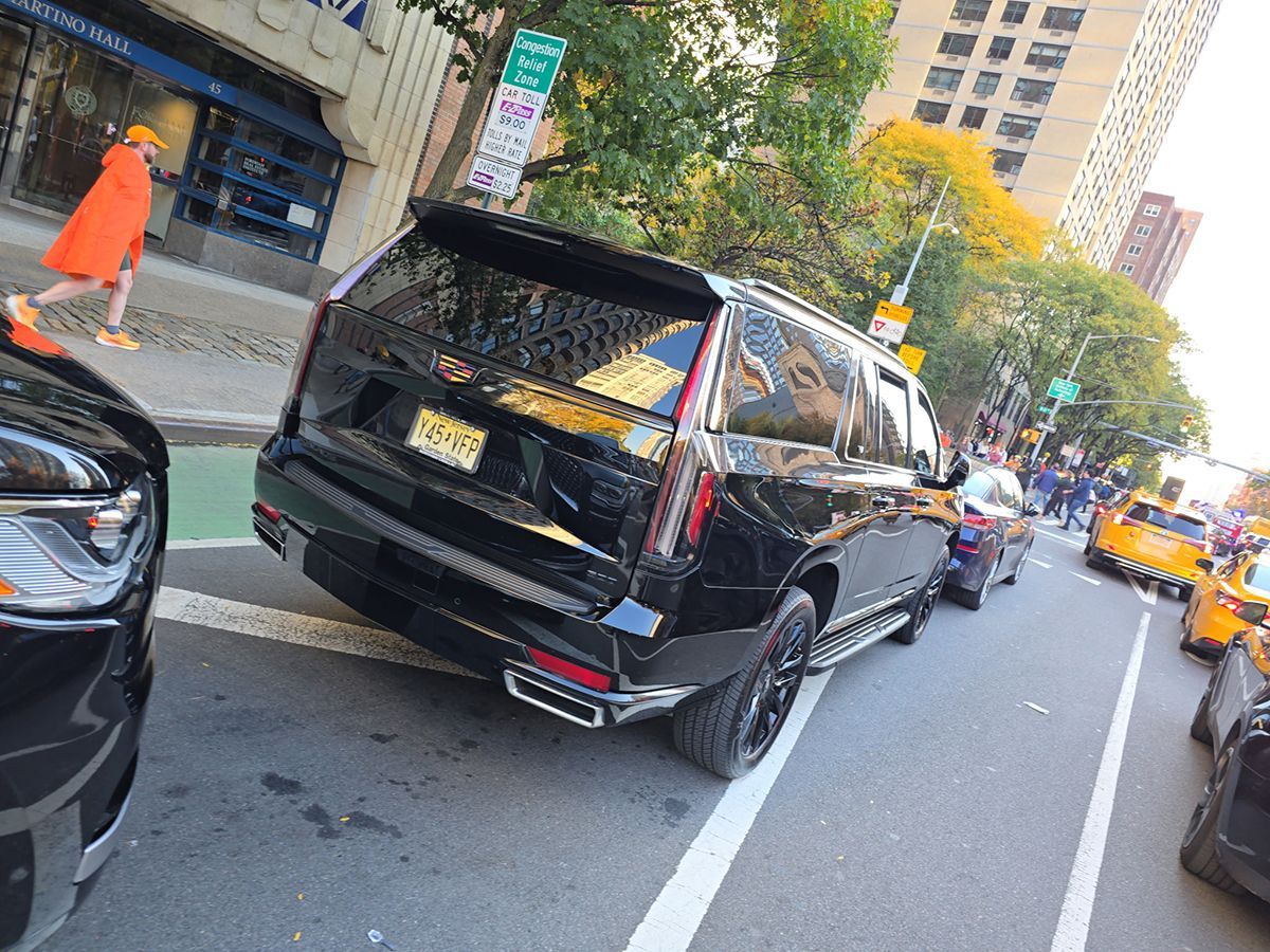 Black Cadillac SUV on city street, person in orange coat walking in background.