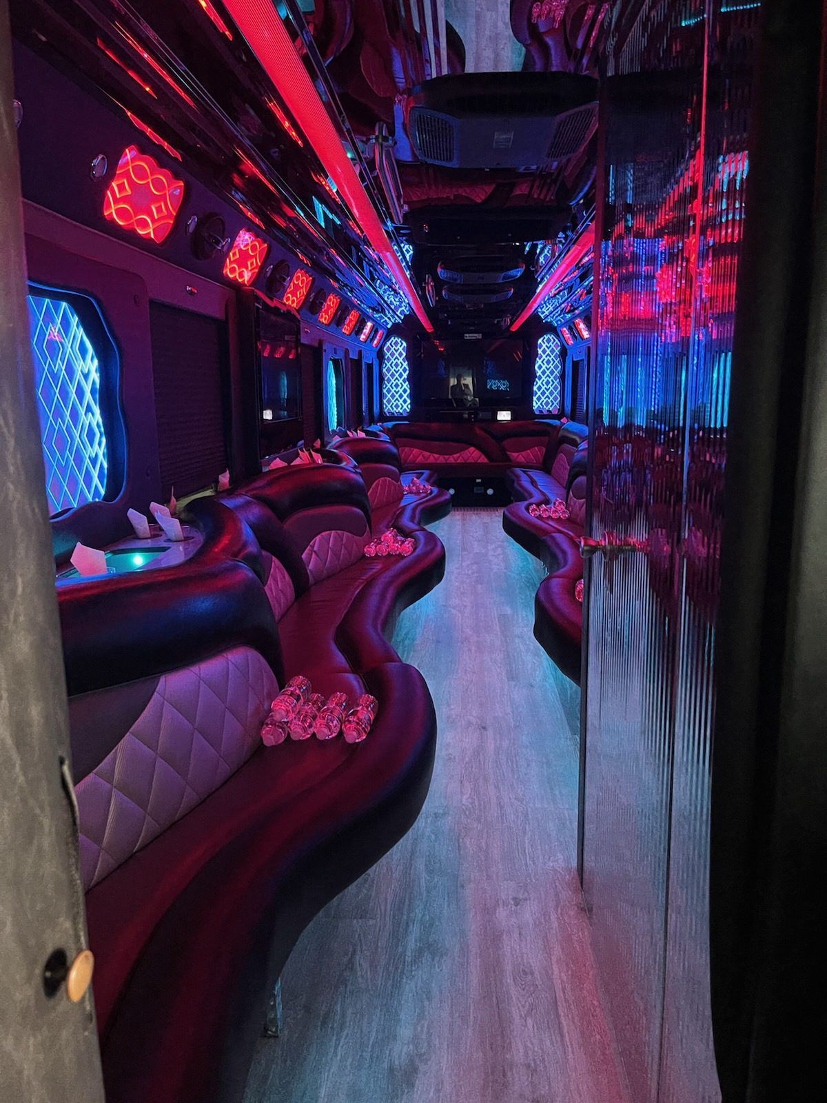 Interior of a party bus with purple and pink LED lights, seating, and a wooden floor.