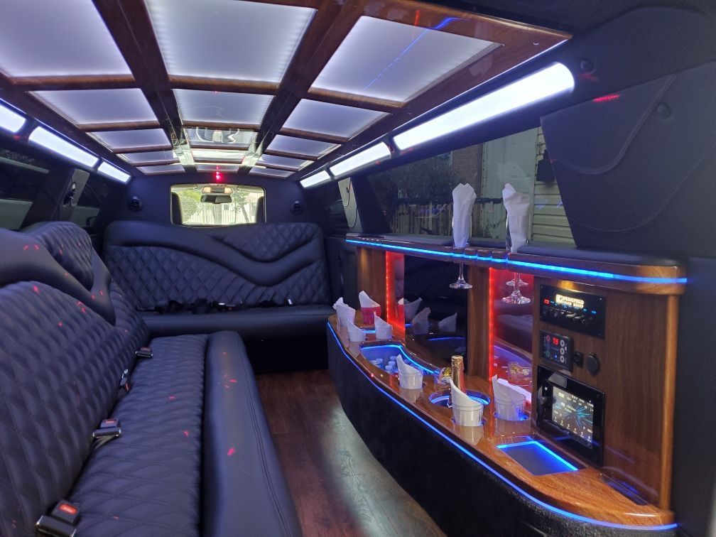 Inside a luxury limousine: black quilted seating, bar, ambient blue lighting, wood trim.