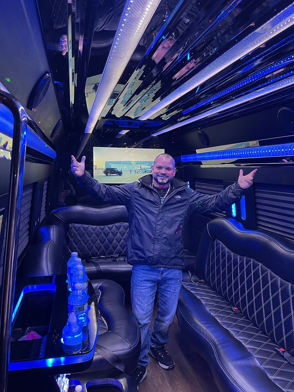 Man in a party bus with arms raised. Blue lights illuminate interior; black seating and accents.