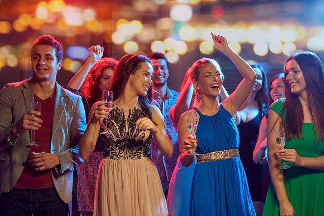 People dancing and celebrating at a party, holding drinks. Bright lights and festive atmosphere.