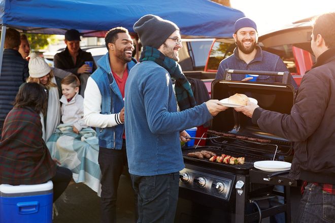 Group of people tailgating at an outdoor event, grilling food, smiling, and socializing.