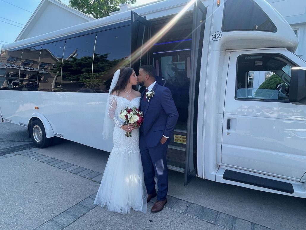 Newlyweds kiss in front of a white party bus. The bride holds a bouquet, the groom wears a blue suit.