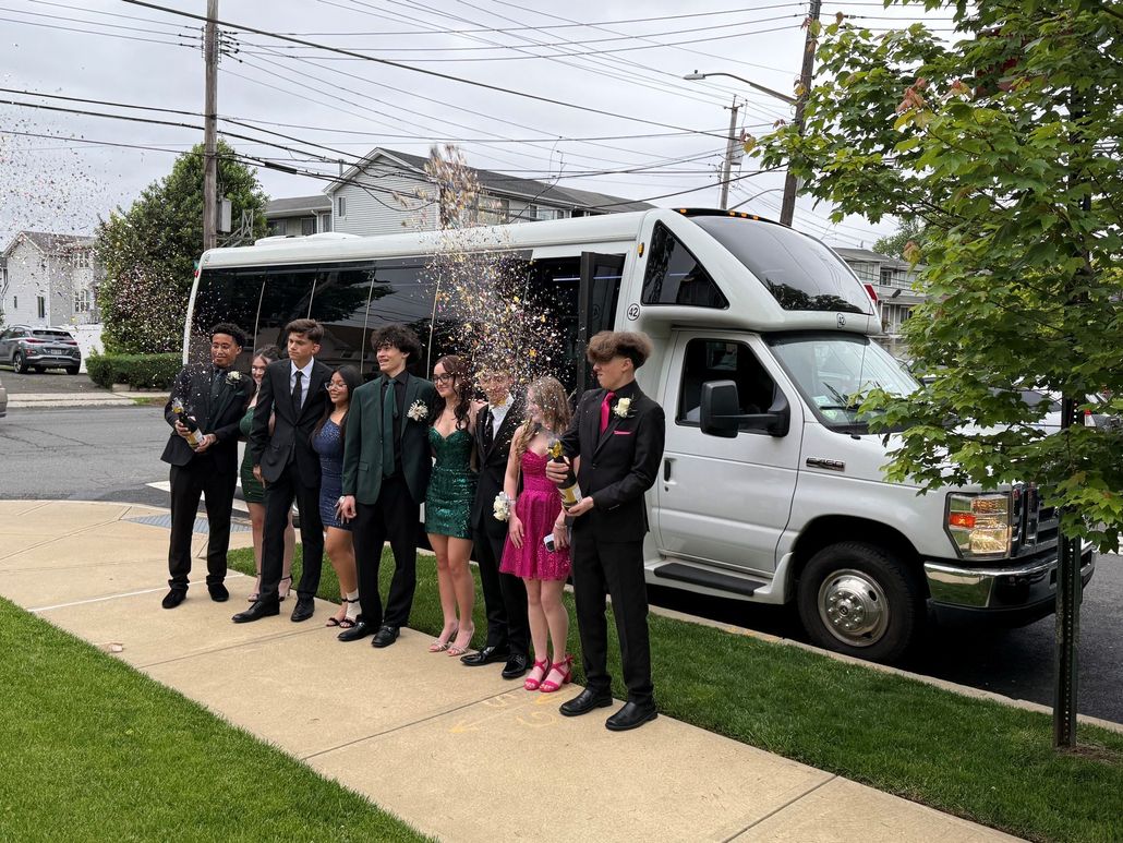 Group of formally dressed teens posing next to a white party bus on a sidewalk. Confetti is in the air.