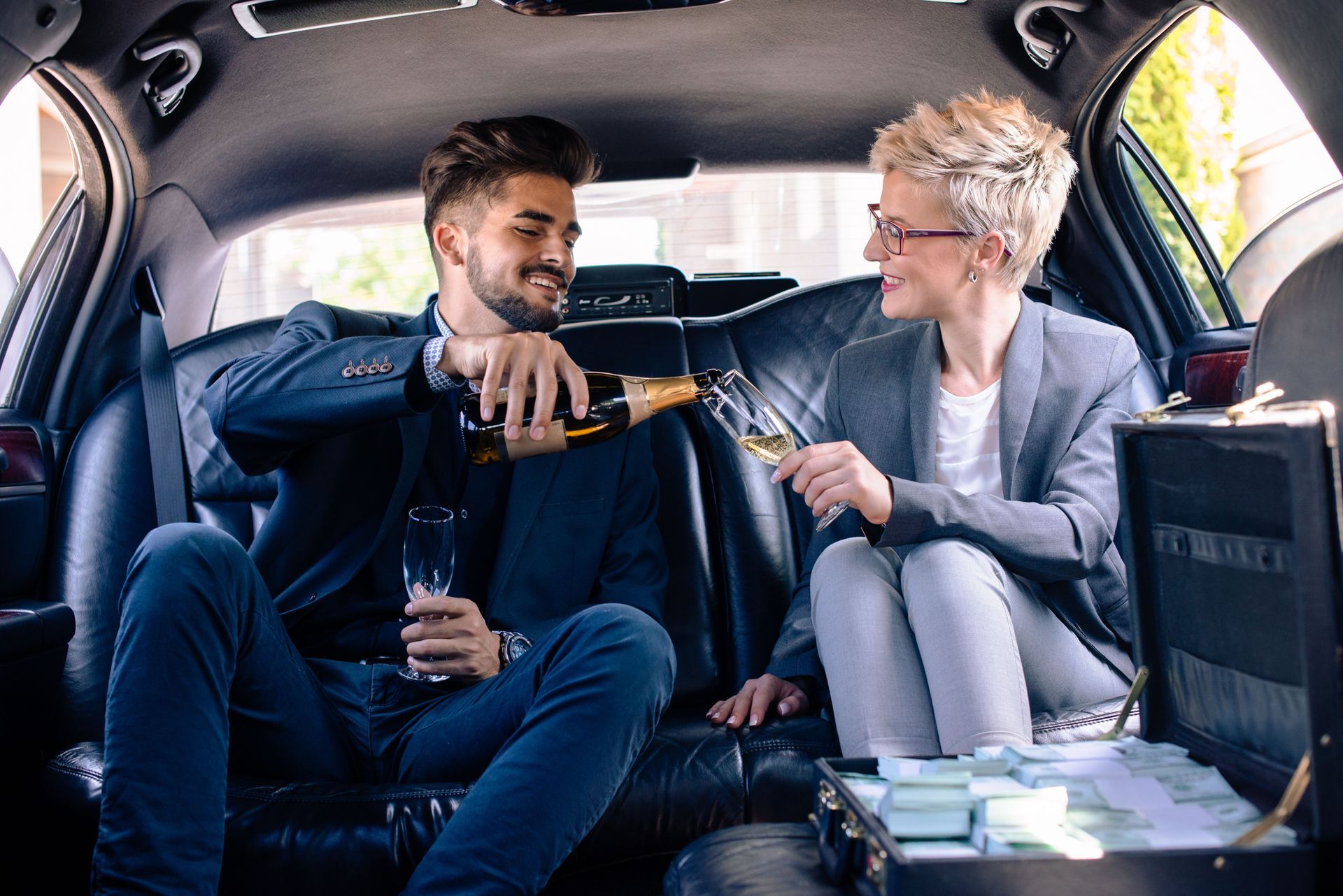 Man pouring champagne for a woman in a limousine; they smile at each other. A briefcase of money is visible.