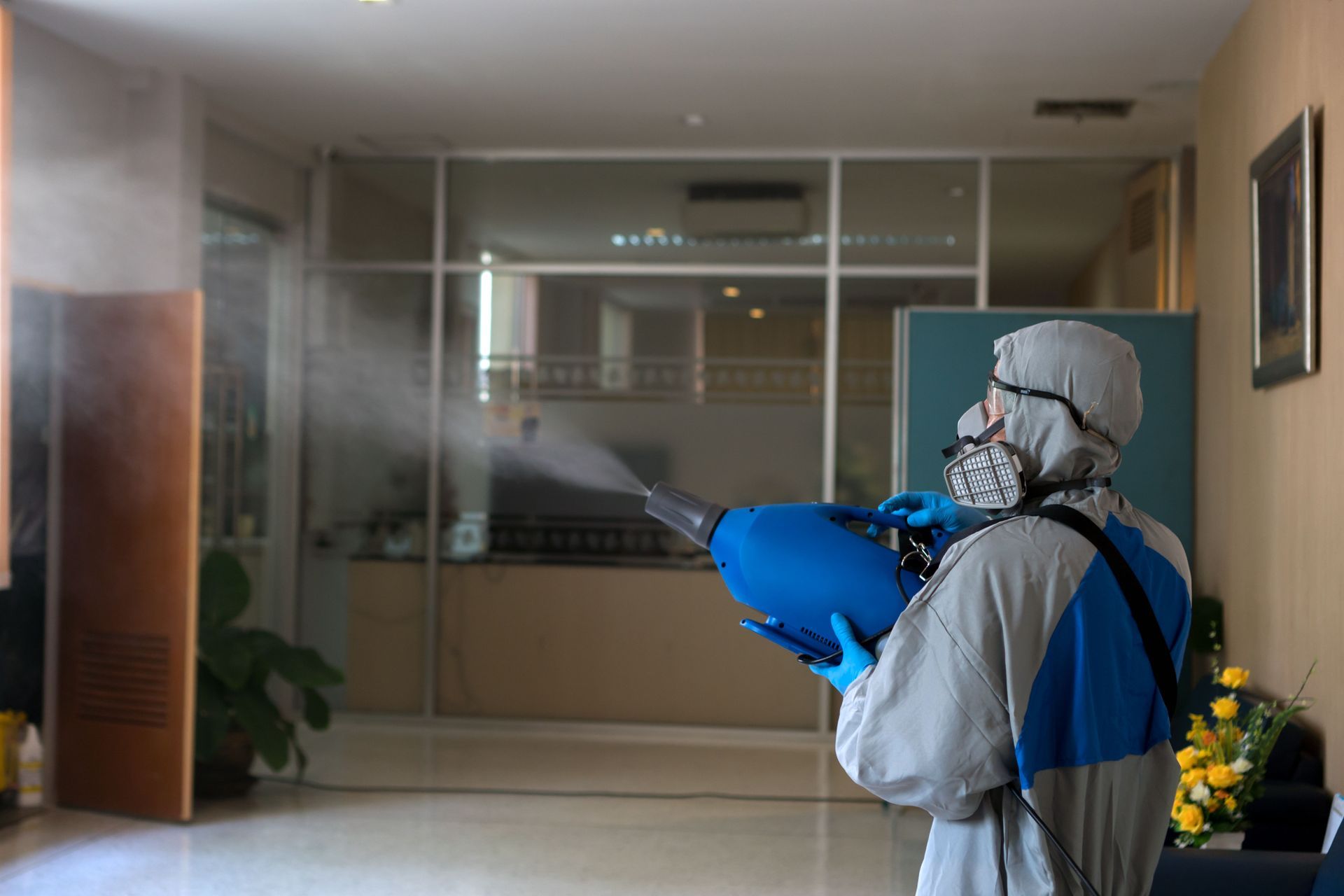 Person in protective gear spraying disinfectant in a hallway with glass doors