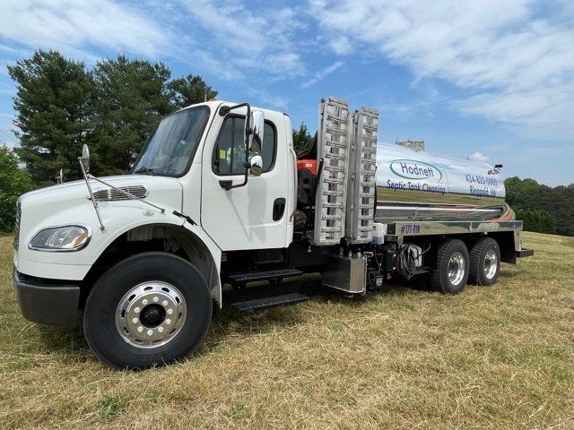 White tanker truck on a grassy field under a blue sky with clouds.