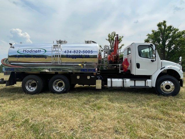 White septic truck with silver tank parked in a field. The company name and phone number are visible.