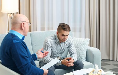 A man is sitting on a couch talking to a man in a living room.