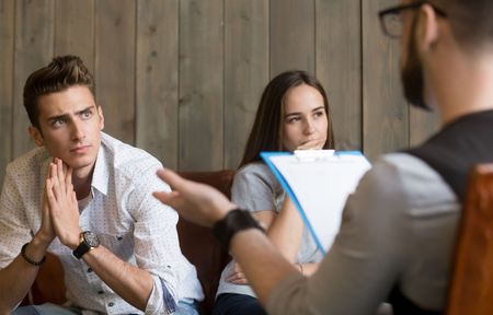 A man and a woman are sitting on a couch talking to a man holding a clipboard.