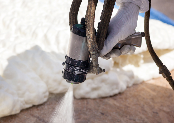 Gloved hand spraying white foam onto a wooden surface outdoors.