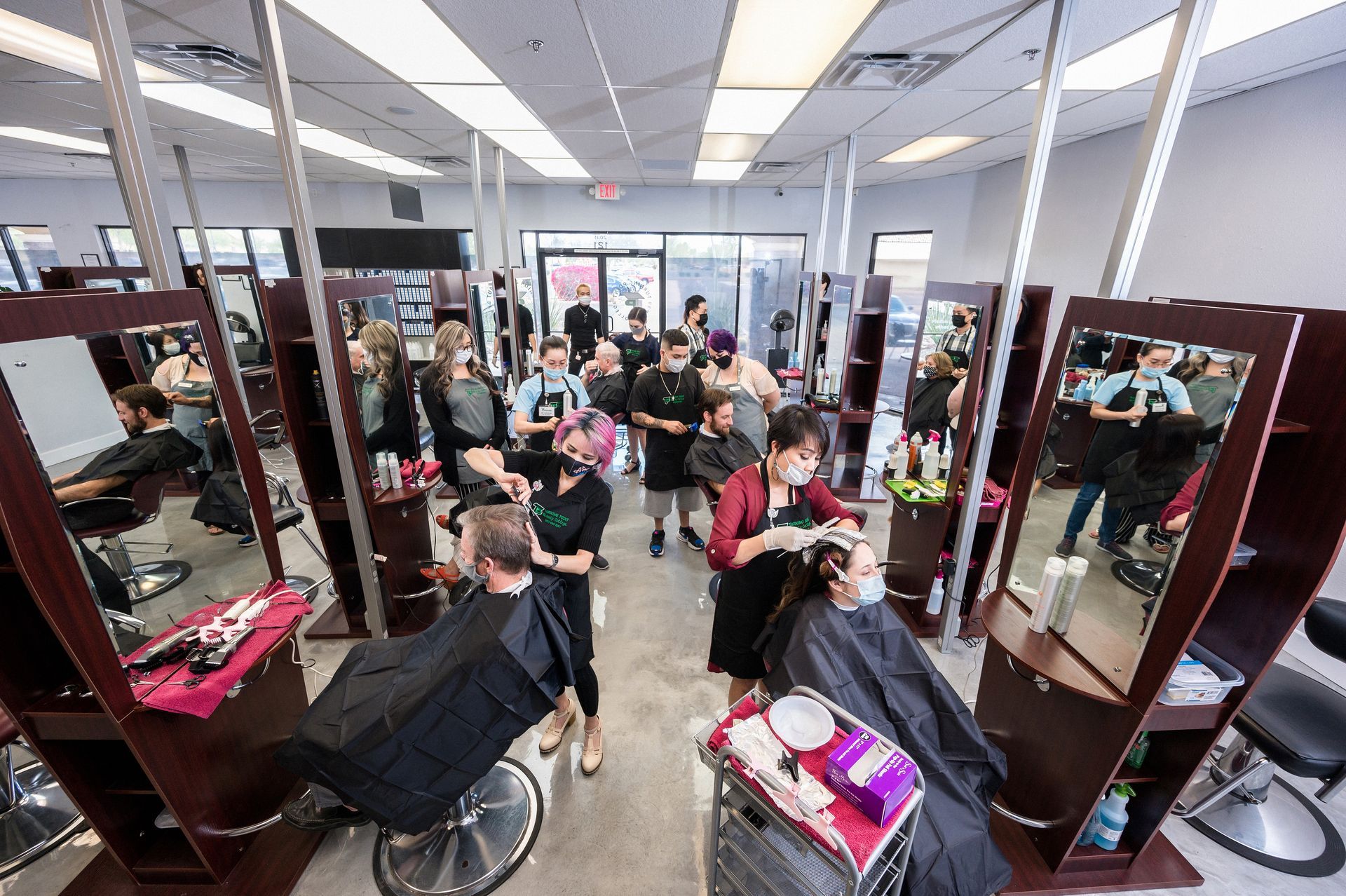 Interior view of a hair salon. Stylists work on clients in front of mirrors. Salon is well-lit, with multiple stations.