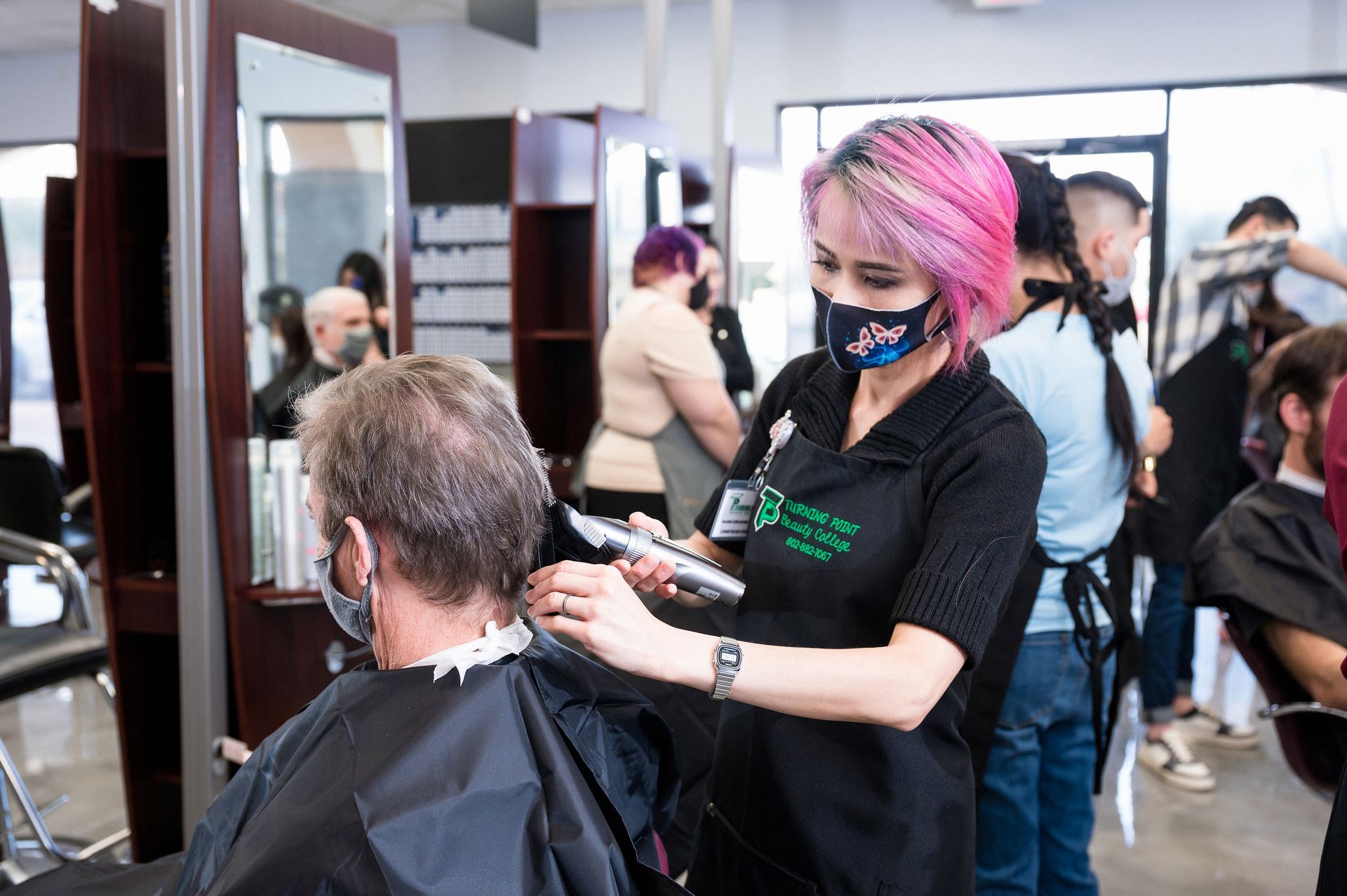 A person with pink hair uses clippers on a client in a salon. Others watch and work.