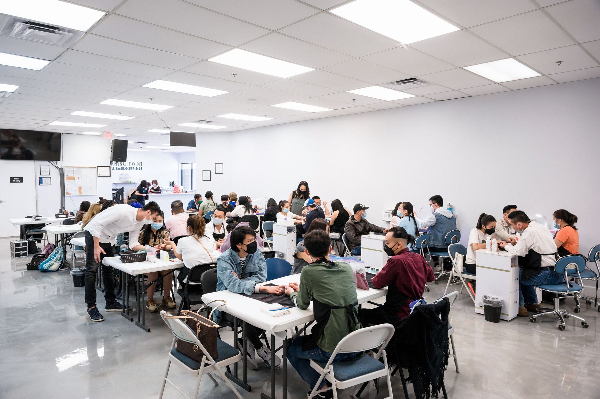A large room with many people, possibly a nail salon training session. People are seated at tables working on nails.