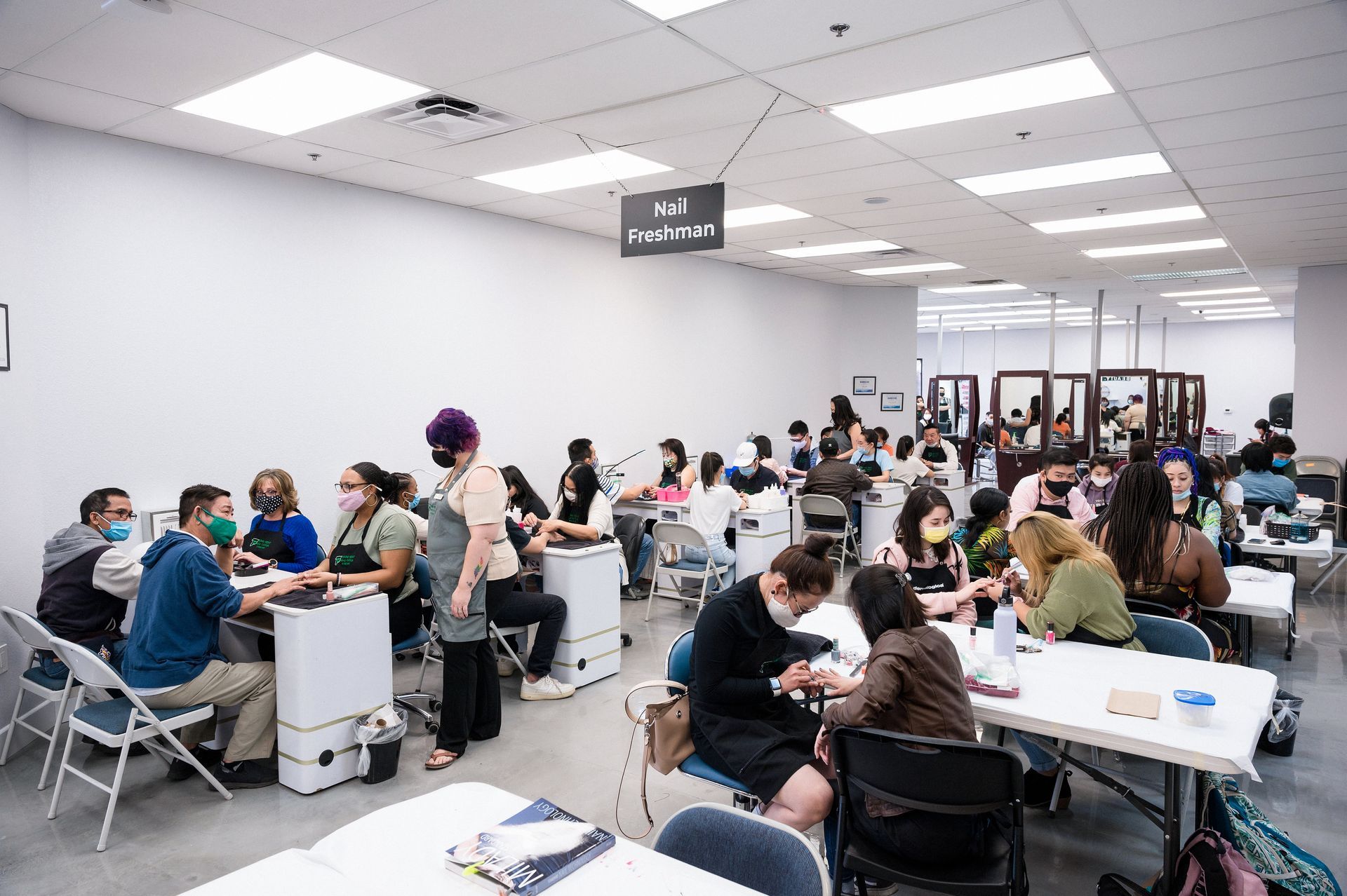 A nail salon with many people sitting at tables, getting manicures. Bright white room with overhead lighting.