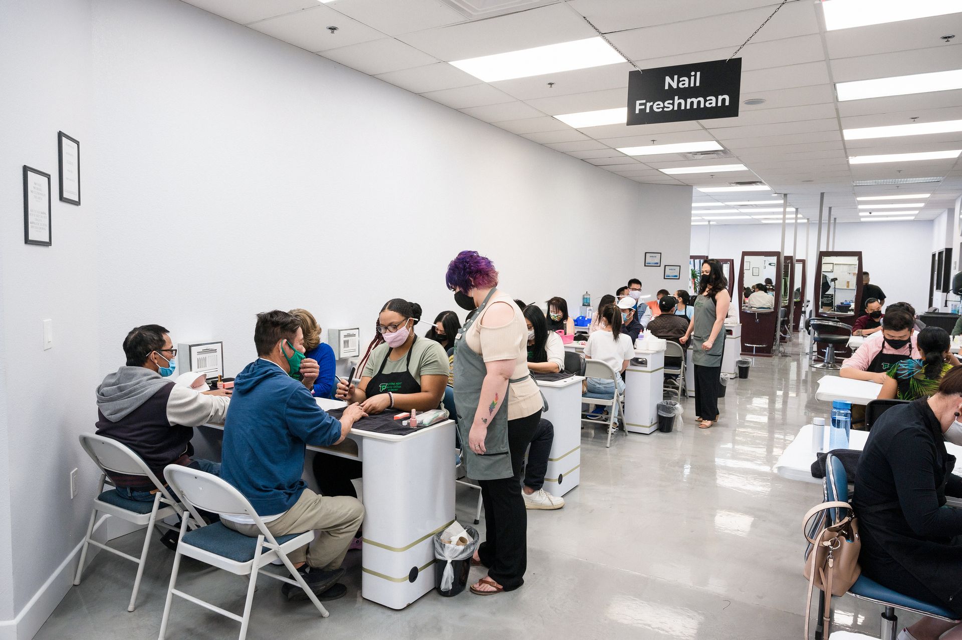 A nail salon with students practicing on clients. White walls, fluorescent lighting, and nail stations.