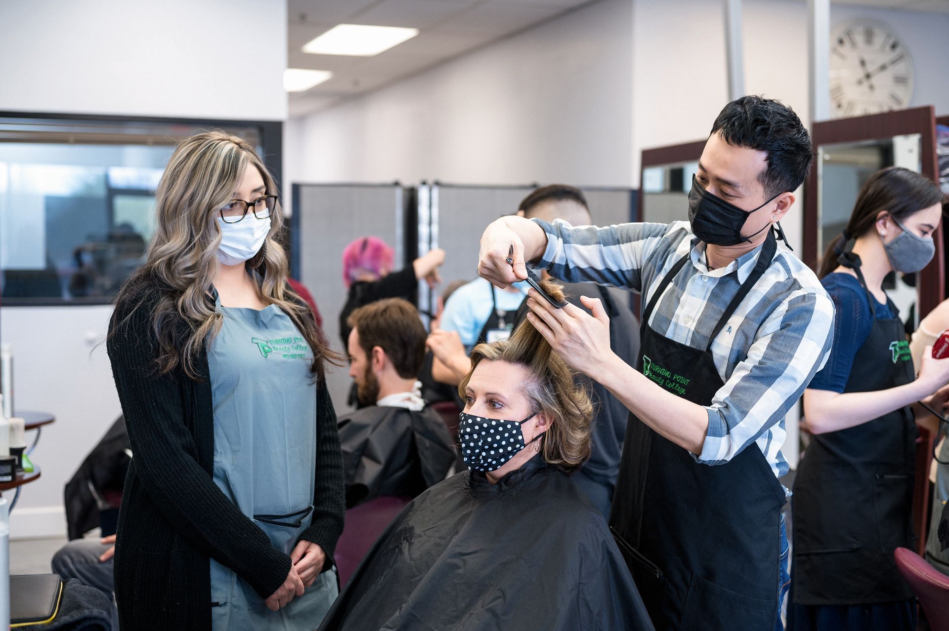 Hair salon: Stylist cutting hair of a client, with other masked students and instructors in the background.