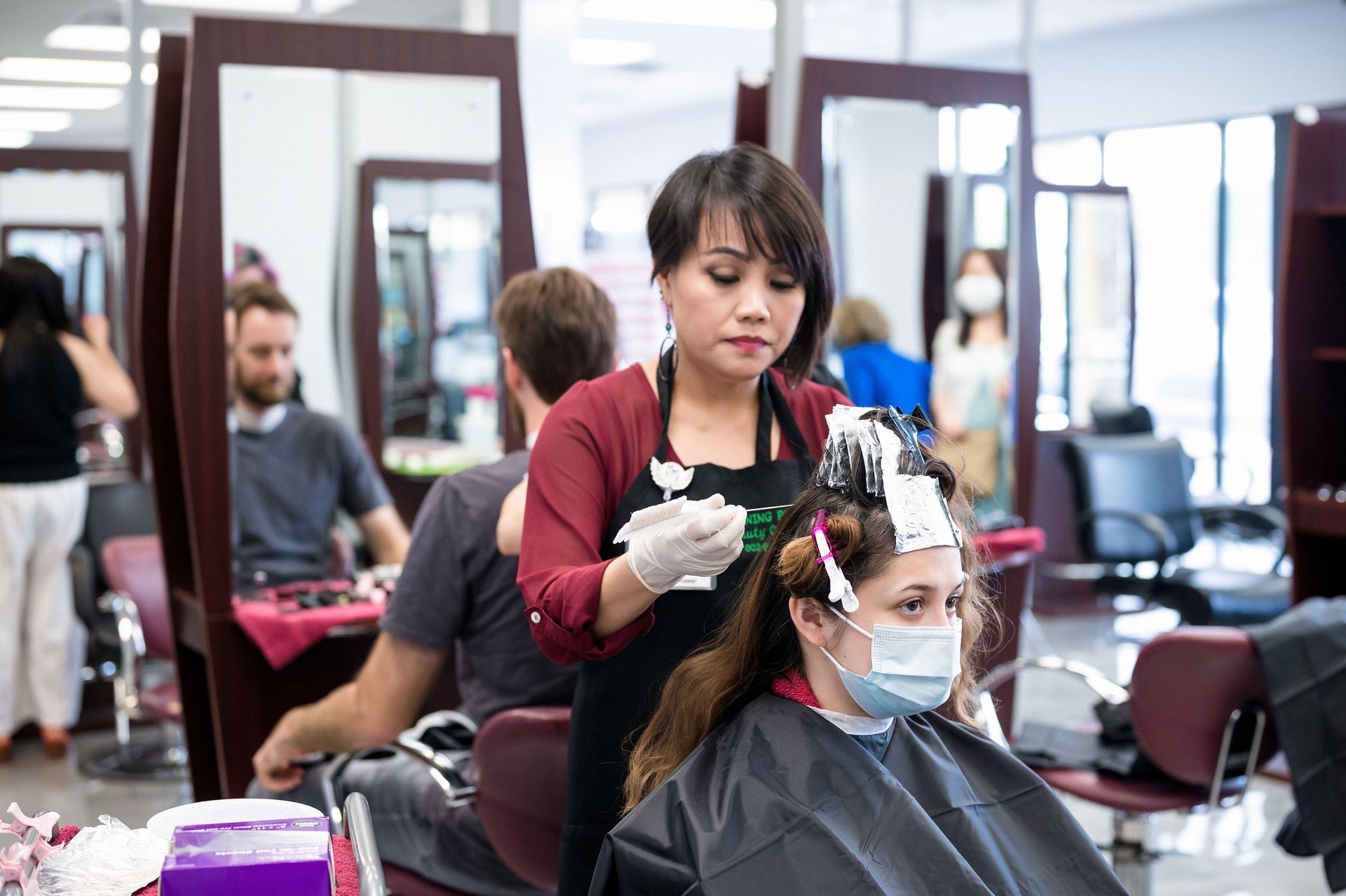 Hairdresser applying hair dye to a customer in a salon. Both wear masks, mirrors and chairs visible.