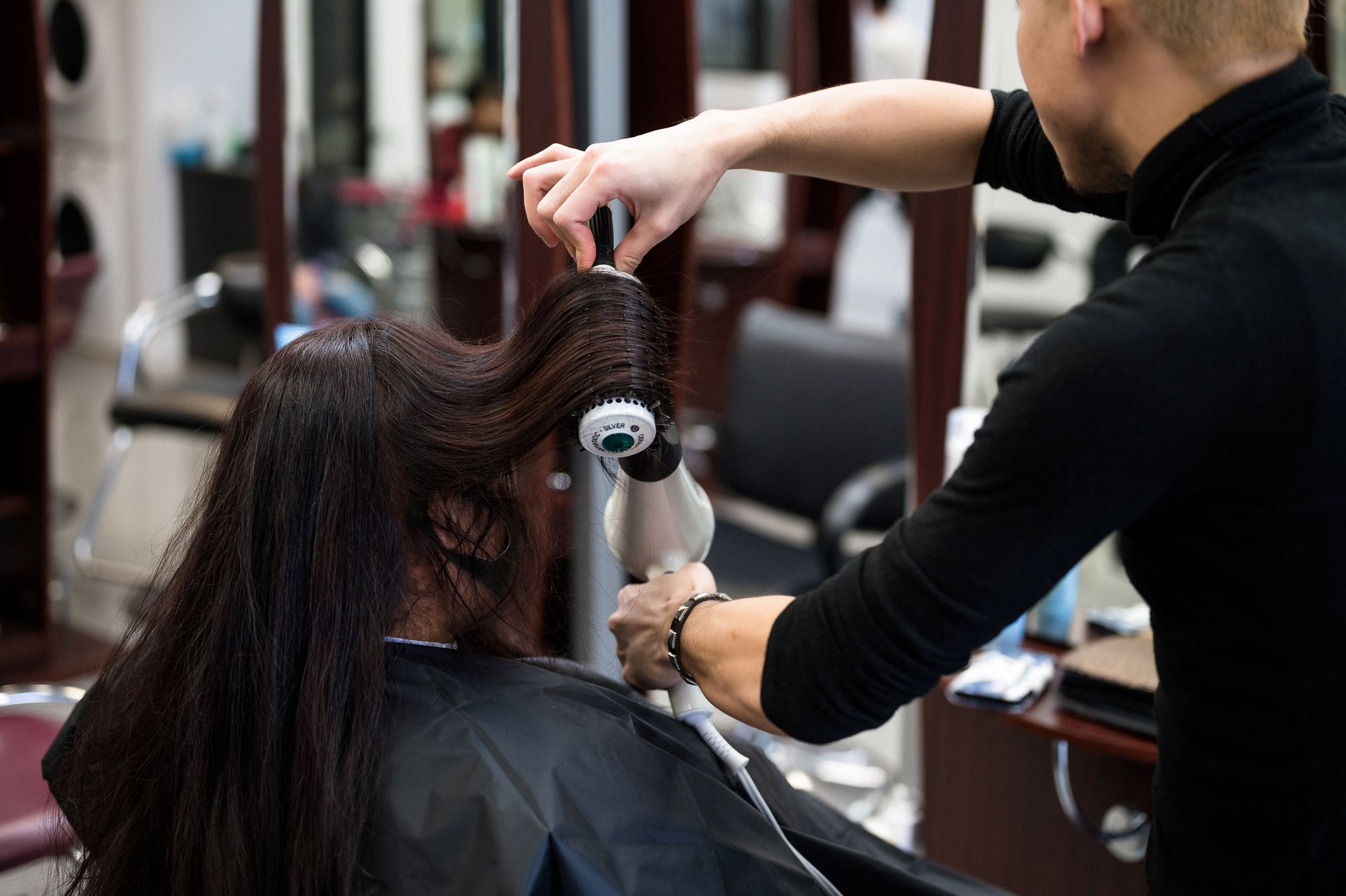 Hair stylist uses a round brush and blow dryer on a client's long, dark hair in a salon.